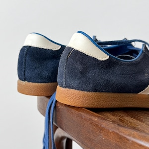 May include: Close-up of navy blue suede sneakers with white and blue accents. The sneakers have brown soles and blue laces. The shoes are placed on a wooden surface.