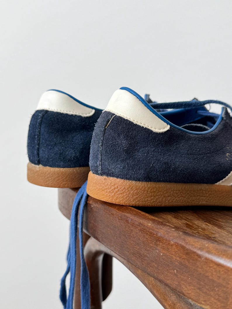 May include: Close-up of navy blue suede sneakers with white and blue accents. The sneakers have brown soles and blue laces. The shoes are placed on a wooden surface.