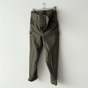 Può includere: Pantaloni cargo verde oliva con più tasche e chiusura con bottoni, appesi a una gruccia di metallo. I pantaloni hanno una vita alta e un tessuto leggermente strutturato. I polsini sono risvoltati. Su sfondo bianco.