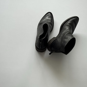 Könnte beinhalten: Ein Paar schwarze Leder-Ankle Boots mit spitzer Zehenpartie und kleinem Absatz. Die Stiefel haben einen Reißverschluss am Rücken und werden auf einer weißen Oberfläche präsentiert.