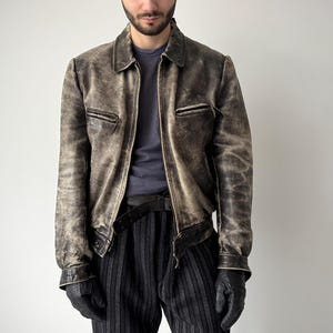 Puede incluir: Chaqueta de cuero marrón desgastado con cierre de cremallera, dos bolsillos en el pecho y cuello. Se lleva sobre una camiseta azul oscuro y se combina con pantalones a rayas negras y guantes negros.