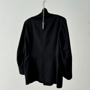 Puede incluir: Una chaqueta negra de traje con un solo bot&oacute;n de cierre y mangas largas. La chaqueta est&aacute; colgada de una cadena plateada.