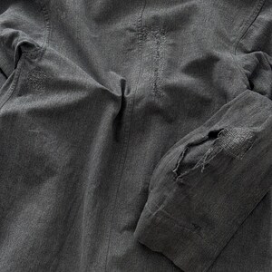 May include: Close-up of a gray jacket with visible distressing and repairs. The jacket features a textured fabric with visible stitching and patches. The elbow of the sleeve has a large patch with visible threadbare areas.