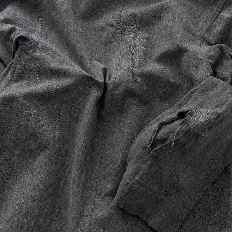 May include: Close-up of a gray jacket with visible distressing and repairs. The jacket features a textured fabric with visible stitching and patches. The elbow of the sleeve has a large patch with visible threadbare areas.