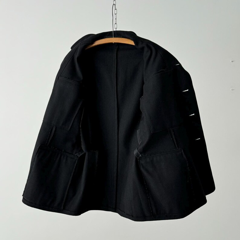 Puede incluir: Una chaqueta negra con cierre de botones y m&uacute;ltiples bolsillos. La chaqueta est&aacute; colgada en una percha de madera.