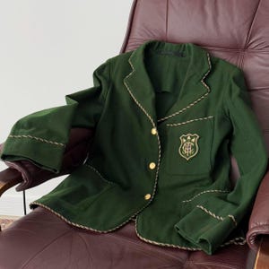 Puede incluir: Una chaqueta verde oscuro con botones y ribetes dorados. La chaqueta tiene un bolsillo en el lado izquierdo con un escudo bordado.