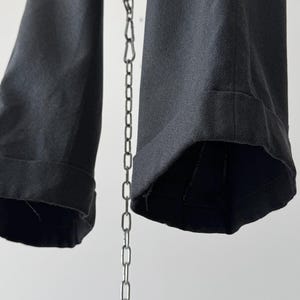 May include: Close-up of dark gray trousers with cuffed hems. The fabric appears to be a woven material. A silver chain hangs in the center, with the trousers suspended from it. The background is a plain white.