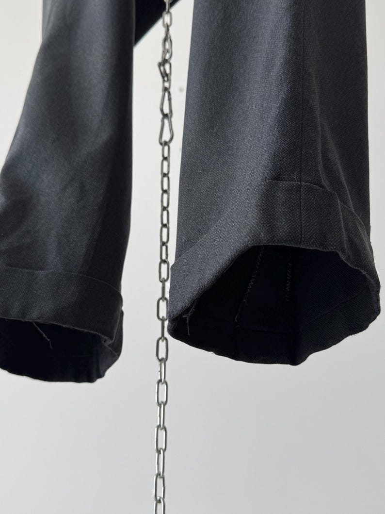 May include: Close-up of dark gray trousers with cuffed hems. The fabric appears to be a woven material. A silver chain hangs in the center, with the trousers suspended from it. The background is a plain white.