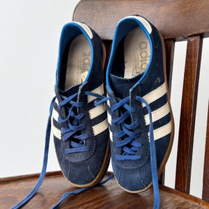 May include: A pair of navy blue sneakers with white stripes and blue laces. The shoes have a suede upper and a brown sole. The sneakers are displayed on a wooden chair.