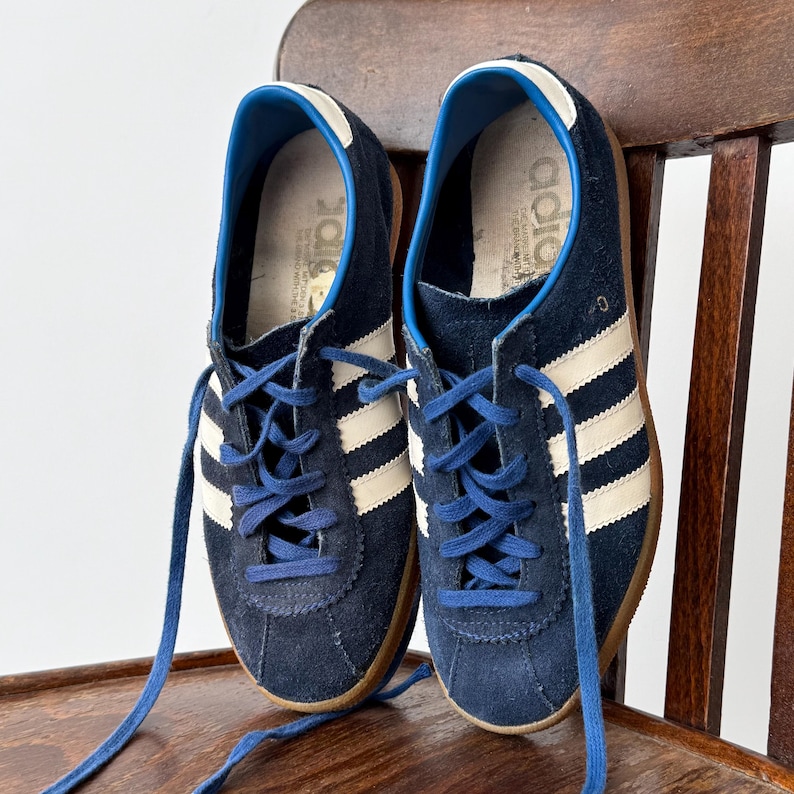 May include: A pair of navy blue sneakers with white stripes and blue laces. The shoes have a suede upper and a brown sole. The sneakers are displayed on a wooden chair.