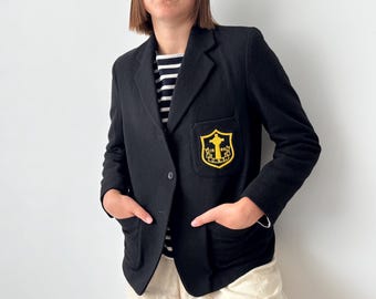 Könnte beinhalten: Schwarzer Blazer mit gelb-schwarzem Wappen auf der Tasche. Der Blazer hat einen Einzelknopfverschluss und zwei Vordertaschen. Die Person trägt ein schwarz-weiß gestreiftes Hemd darunter.