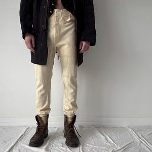 Könnte beinhalten: Cremefarbene Hose mit hohem Bund und Knopfleiste, kombiniert mit braunen Lederstiefeln. Eine schwarze Jacke wird offen getragen und zeigt die Hose. Die Hose hat eine helle, neutrale Farbe, und die Stiefel haben Schnürsenkel.