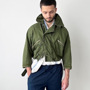 Może przedstawiać: Oliwkowozielona krótka kurtka z dużym kołnierzem, dwiema kieszeniami na zamek i zapięciem na guziki. Kurtka jest noszona na białej koszuli w niebieskie paski i granatowych spodniach. Kurtka ma pasek.