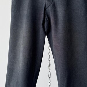 May include: Close-up of black dress pants with a straight leg design. The pants are made of a smooth fabric and have a classic, tailored look. A silver chain hangs from the pants.