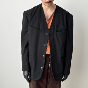 Puede incluir: Un blazer negro con diseño de cuello en V, con tres botones y un bolsillo en el lado izquierdo. El blazer se usa sobre una camisa naranja y pantalones marrones. La persona lleva guantes negros.