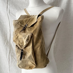 May include: A tan canvas backpack with a flap closure and a front pocket. The backpack has adjustable shoulder straps and leather accents. The bag is displayed on a white mannequin.