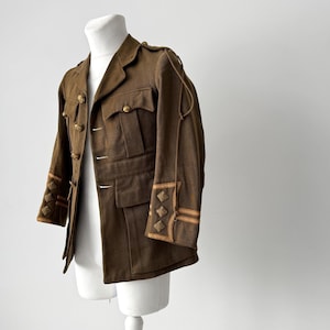 Może przedstawiać: Brązowa kurtka wojskowa ze złotymi guzikami i dekoracyjnymi akcentami na rękawach. Kurtka, prezentowana na manekinie, ma wiele kieszeni. Odzież wojskowa w stylu vintage.