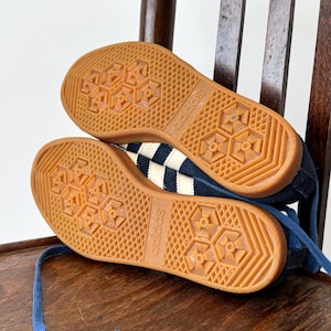 May include: A pair of navy blue Adidas sneakers with white stripes and a gum rubber sole. The soles are visible, showcasing the Adidas logo and a textured pattern. The laces are blue.