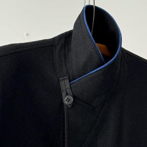 Puede incluir: Una chaqueta negra con ribete azul en el cuello. La chaqueta tiene un cierre de bot&oacute;n &uacute;nico y un detalle de dise&ntilde;o &uacute;nico en la parte delantera.
