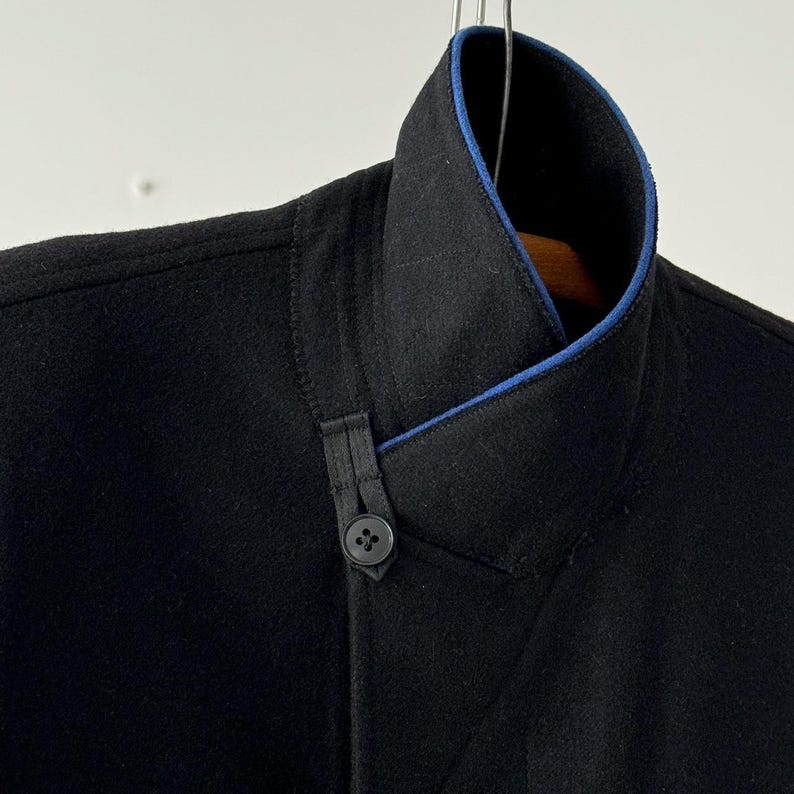 Puede incluir: Una chaqueta negra con ribete azul en el cuello. La chaqueta tiene un cierre de bot&oacute;n &uacute;nico y un detalle de dise&ntilde;o &uacute;nico en la parte delantera.