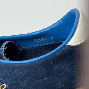 May include: Close-up of a navy blue suede sneaker interior. The shoe features a blue leather collar with a white accent. The number "31560" and the letter "E" are printed in gold. The sole is a light brown color.