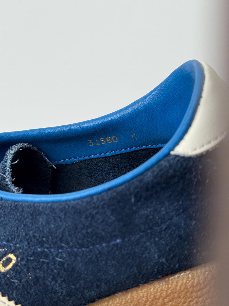 May include: Close-up of a navy blue suede sneaker interior. The shoe features a blue leather collar with a white accent. The number "31560" and the letter "E" are printed in gold. The sole is a light brown color.