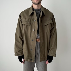 Puede incluir: Chaqueta abotonada verde oliva con dos bolsillos en el pecho. La chaqueta se lleva sobre una camiseta gris y pantalones grises. El modelo también lleva guantes negros.