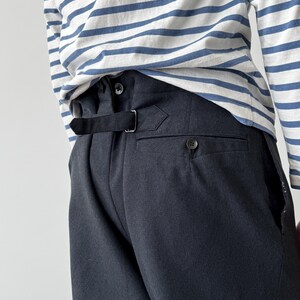 May include: Close-up of the back of dark gray trousers with a black belt and button detail. The trousers are paired with a white and blue striped long-sleeve shirt. The fabric appears to be a woven material.