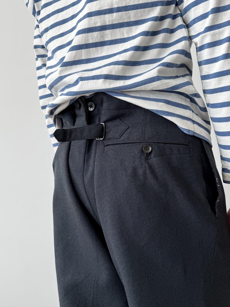 May include: Close-up of the back of dark gray trousers with a black belt and button detail. The trousers are paired with a white and blue striped long-sleeve shirt. The fabric appears to be a woven material.
