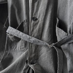 May include: Close-up of a gray denim jacket with a matching belt. The jacket features a button-down front, two front pockets, and a belt that cinches at the waist. The fabric has a textured appearance.