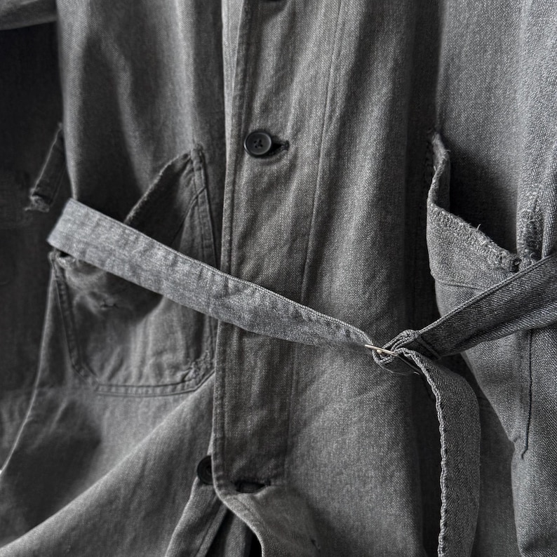 May include: Close-up of a gray denim jacket with a matching belt. The jacket features a button-down front, two front pockets, and a belt that cinches at the waist. The fabric has a textured appearance.