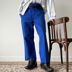 Può includere: Pantaloni blu brillante con cintura nera. I pantaloni sono abbinati a scarpe nere e una camicia a righe. Una sedia di legno è sullo sfondo. I pantaloni sono di un colore vibrante e sembrano essere fatti di un materiale resistente.