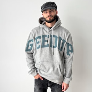 Może przedstawiać: Szara bluza z kapturem z napisem "GEEDUP" w turkusowych literach na piersi. Bluza ma kaptur ze sznurkiem i przednią kieszeń. Osoba na zdjęciu ma na sobie ciemną czapkę i ciemne dżinsy.