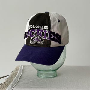 May include: A vintage baseball cap in a black, white and purple colour scheme. The front of the cap features the words "Colorado Rockies" in a purple and white embroidered design. The cap has a navy blue brim.