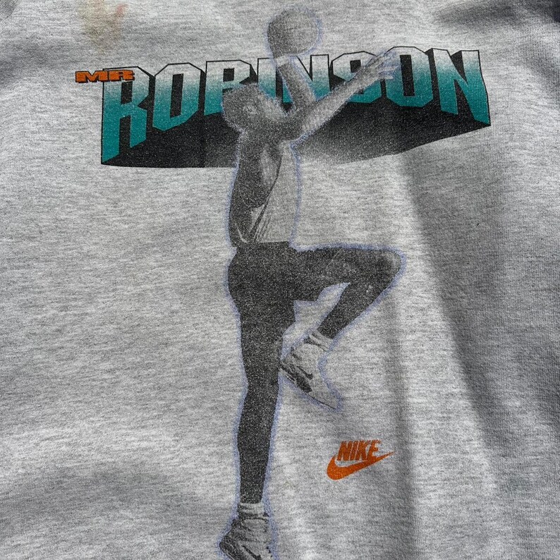 Puede incluir: Camiseta gris con un gr&aacute;fico de un jugador de baloncesto en el aire, con el texto "Mr. Robinson" sobre el jugador y un logotipo de Nike debajo.