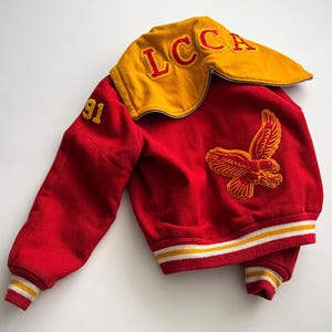 Op de afbeelding: Een vintage rode en gouden letterman-jas met de letters "LCCA" op de kraag en het nummer "91" op de mouw. De jas heeft een geborduurde adelaar op de voorkant en gestreepte manchetten.