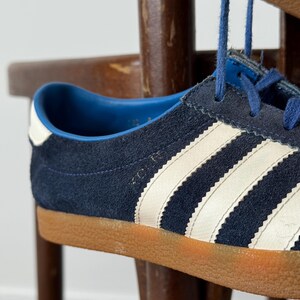 May include: Close-up of a navy blue suede sneaker with white stripes and a brown sole. The shoe has blue laces and a white heel accent. The sneaker is a classic athletic style.