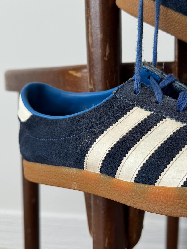 May include: Close-up of a navy blue suede sneaker with white stripes and a brown sole. The shoe has blue laces and a white heel accent. The sneaker is a classic athletic style.