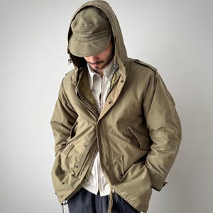 以下が含まれることがあります： オリーブグリーンのフード付きジャケットと、お揃いのキャップ。 ジャケットはジッパー開閉、複数のポケット、ウエストにドローコードが付いています。 下には白い襟付きシャツが着用されています。