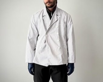 May include: A double-breasted jacket featuring a white and navy blue pinstripe pattern, a notched lapel, and four white buttons on the front. The jacket includes long sleeves and a chest pocket, worn over a dark gray zip-up shirt and black pants.