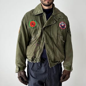 May include: Olive green military-style jacket with patches. The jacket has a collared neckline, multiple pockets, and a button-up front. Patches include text that reads "Maritime Strike Attack". The jacket is worn with a navy blue sweater and gray pinstripe pants.