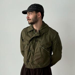 Puede incluir: Chaqueta verde oliva de estilo militar con cierre de botones, dos bolsillos con cremallera y cuello. La chaqueta se combina con una gorra negra. El diseño de la chaqueta sugiere una estética vintage o retro.