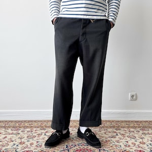 May include: Black trousers with a cuffed hem, paired with black loafers. The person is wearing a striped long-sleeve shirt. The outfit is set against a white wall and a patterned rug.