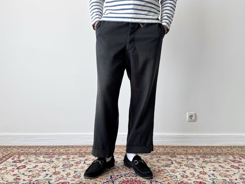 May include: Black trousers with a cuffed hem, paired with black loafers. The person is wearing a striped long-sleeve shirt. The outfit is set against a white wall and a patterned rug.