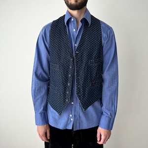 Puede incluir: Un chaleco azul marino con un pequeño estampado de lunares blancos sobre una camisa abotonada azul claro. El chaleco tiene cierre de cinco botones y dos bolsillos delanteros. La persona lleva pantalones negros.