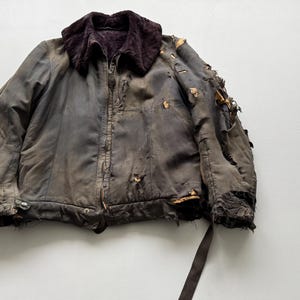 Op de afbeelding: Een versleten vintage jas in grijze en bruine tinten. Het heeft een donkerbruine, pluche kraag en een ritssluiting. De stof vertoont aanzienlijke slijtage, met gaten en gerafelde randen, waardoor het een versleten uiterlijk heeft.