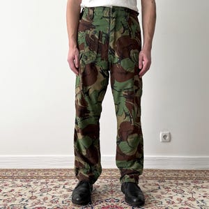 Könnte beinhalten: Camouflagehose mit einem grün-braun-schwarzen Muster. Die Hose hat eine hohe Taille und ein gerades Bein.