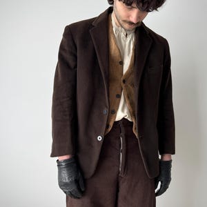 May include: A dark brown corduroy blazer, vest, and trousers. The blazer has two buttons and a pocket. The vest is a lighter brown. The trousers are high-waisted with a zip. The person is wearing black gloves. The outfit has a vintage aesthetic.