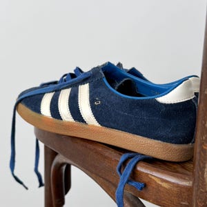 May include: Navy blue suede sneakers with white stripes and a brown sole. The sneakers have blue laces and a white heel tab. The shoes are resting on a wooden chair.