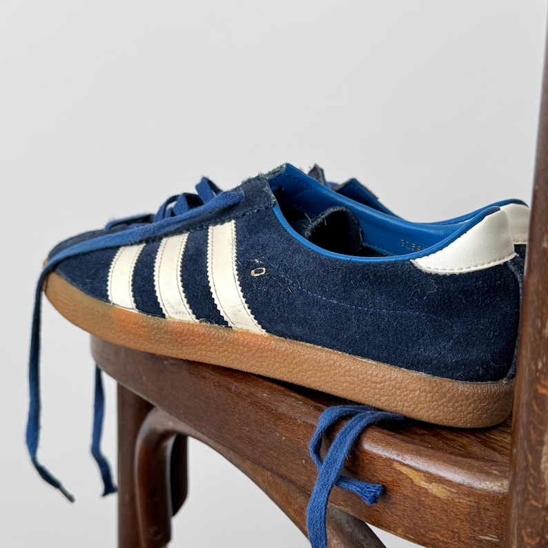 May include: Navy blue suede sneakers with white stripes and a brown sole. The sneakers have blue laces and a white heel tab. The shoes are resting on a wooden chair.